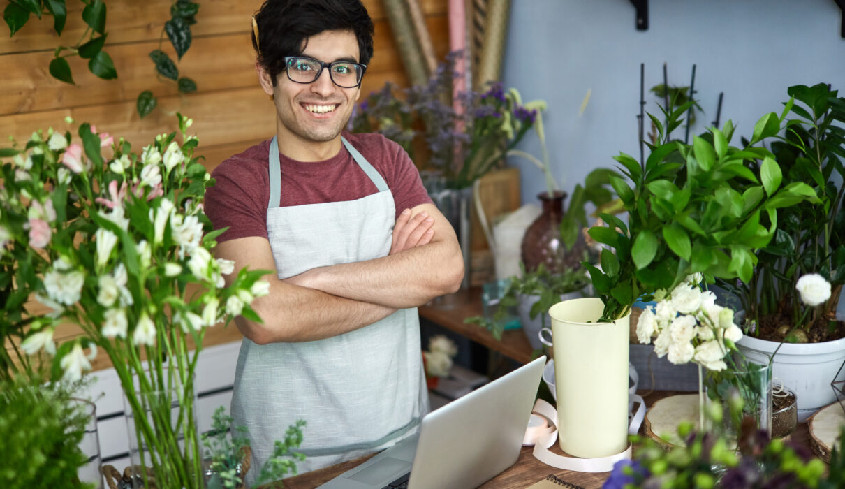 vendedor usando óculos, avental, com braços cruzados em uma empresa de pequeno porte de venda de flores