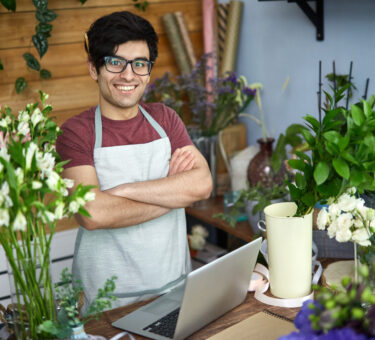 vendedor usando óculos, avental, com braços cruzados em uma empresa de pequeno porte de venda de flores