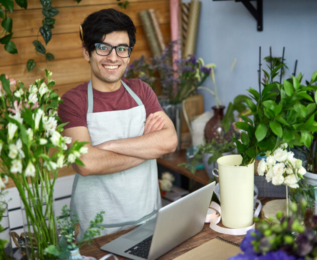 vendedor usando óculos, avental, com braços cruzados em uma empresa de pequeno porte de venda de flores